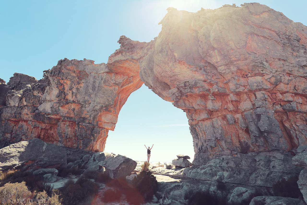 Cederberg's Celestial Arch Foto von A lone traveler stands beneath the majestic rock arch in Cederberg, South Africa, basking in sunlight and nature's grandeur.